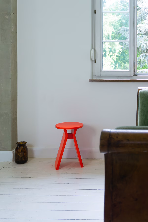 Hocker „Koshi“ in modernem Interieur Ein leuchtend roter Hocker mit dem Namen "Koshi" steht in einer minimalistisch eingerichteten Ecke eines Raumes mit weißem Holzboden. Daneben eine braune Vase, im Hintergrund ein Fenster mit Blick ins Grüne und ein Teil eines rustikalen Sofas.