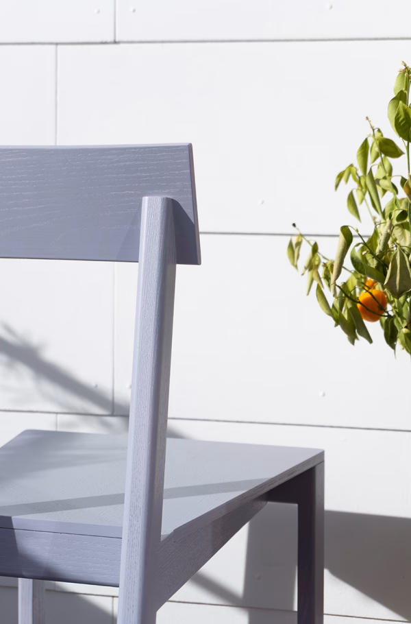 Stuhl „Ari“ in sanftem Grauton im Sonnenlicht Eine Nahaufnahme des grauen Holzstuhls "Ari" vor einer weißen Wand. Sonnenlicht wirft Schatten auf die Oberfläche, während ein Orangenbaum mit leuchtenden Früchten ins Bild ragt.