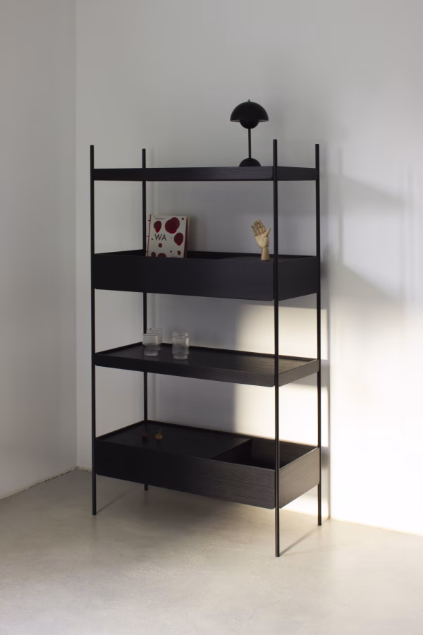 Minimalistisches Regal „Felo“ von Wolter Furniture Das schwarze Metallregal "Felo" von Wolter Furniture steht in einem schlichten, hellen Raum. Es hat vier Ablagen, auf denen sich eine kleine Lampe, ein Buch mit roten Punkten, eine Holzhand, zwei Wassergläser und dekorative Kreisel befinden.